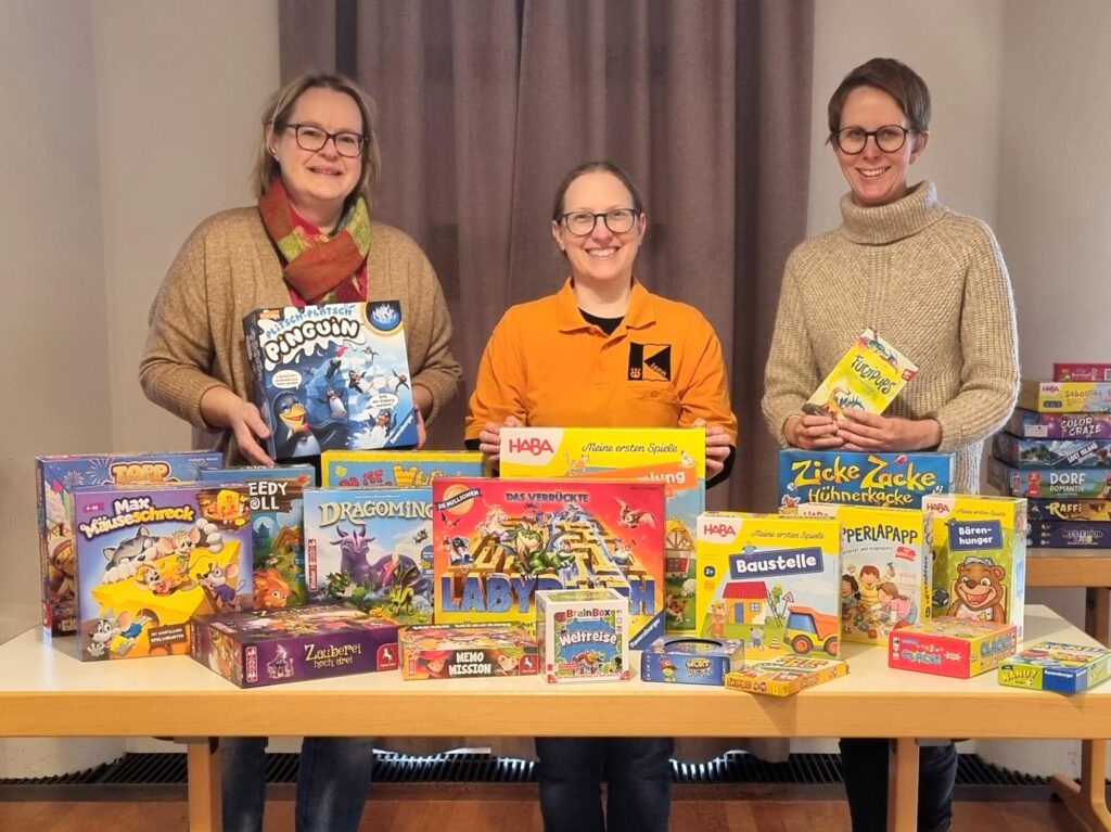 Das Bild zeigt Kerstin Aicher (links) und Franziska Hartmann (rechts) von der Bücherei sowie Annegret Scholz von der Kolpingsfamilie Isen hinter einem Tisch mit Kinderspielen.